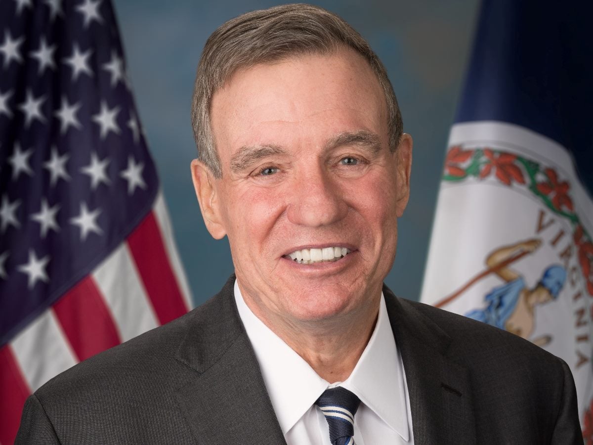 U.S. Senator Mark Warner of Virginia poses for an official portrait, wearing a dark suit and striped tie, with the American and Virginia state flags in the background.