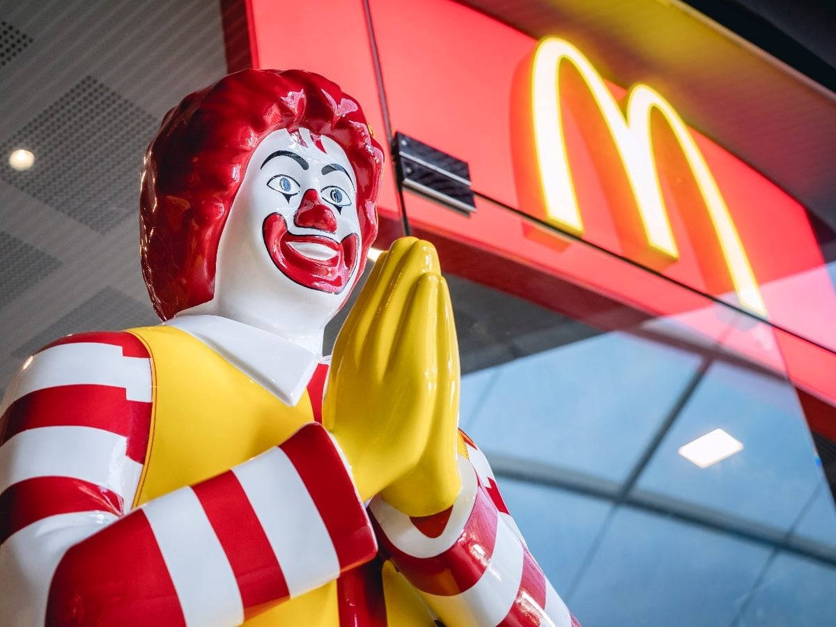 Ronald McDonald statue with hands clasped in front of a McDonald’s restaurant, with the illuminated golden arches sign visible in the background.
