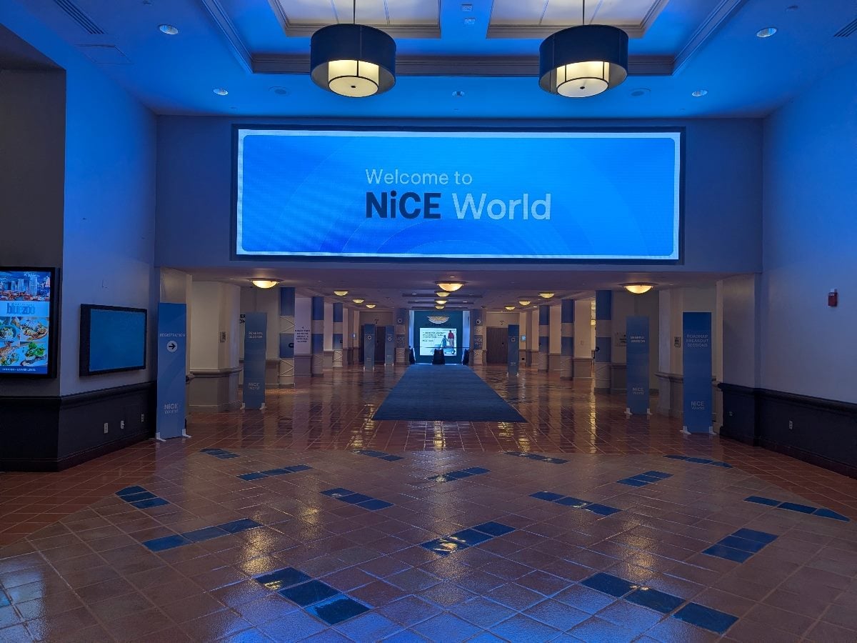 Wide-angle photograph of the entrance concourse at NiCE World 2026 inside the Walt Disney Swan and Dolphin Resort in Orlando. A large illuminated blue digital sign reading “Welcome to NiCE World” hangs above the main hallway, while blue event wayfinding signs line both sides of the corridor. The tiled floor reflects the blue lighting from the display, creating a futuristic atmosphere that mirrors the conference's focus on AI and customer experience innovation.