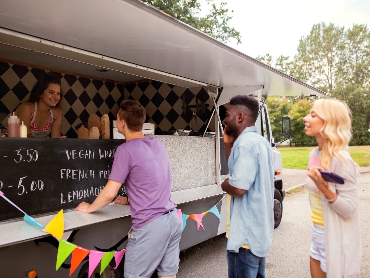 Customers line up at a food truck in a park setting while a staff member takes orders from the service window. Three customers wait at the counter, with menu items and prices displayed on a chalkboard sign. The scene illustrates a simple, in-person customer experience where convenience, speed and ease of service influence satisfaction and loyalty.