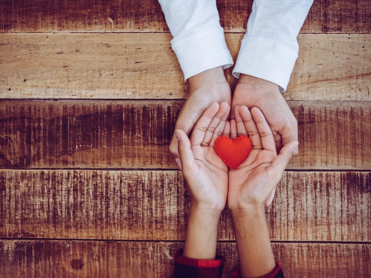 Two pairs of hands gently holding a small red heart-shaped object together over a rustic wooden surface, symbolizing care, compassion, support or human connection.