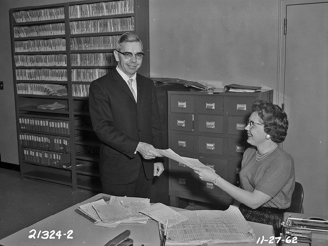 Engineering Department employees for the city of Seattle, 1962. A standing man takes a piece of paper from a sitting woman.