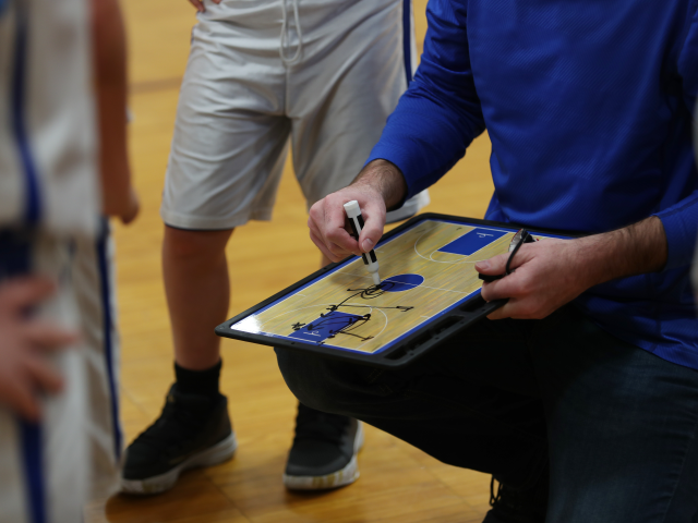 A basketball coach in a team huddle of players writes a play on the playbook.