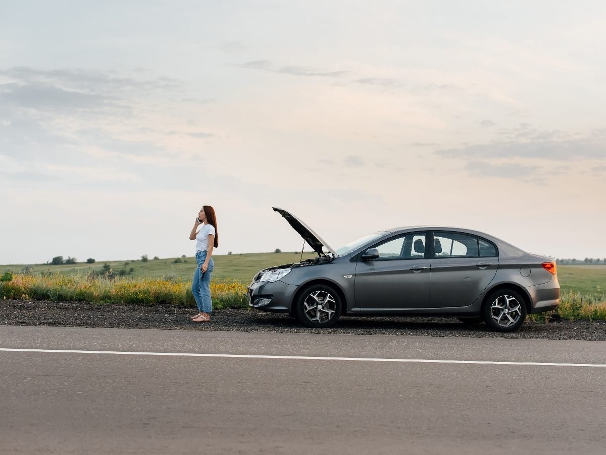 Woman standing on the roadside talking on the phone next to a gray car with its hood open, stranded in a rural area.