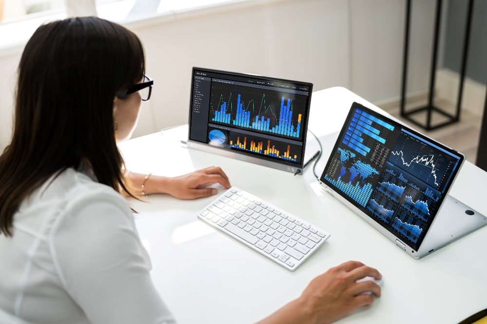 A person with dark hair and wearing glasses and a white shirt sits at a desk viewing two computer screens displaying analytics dashboards in piece about digital customer experience.