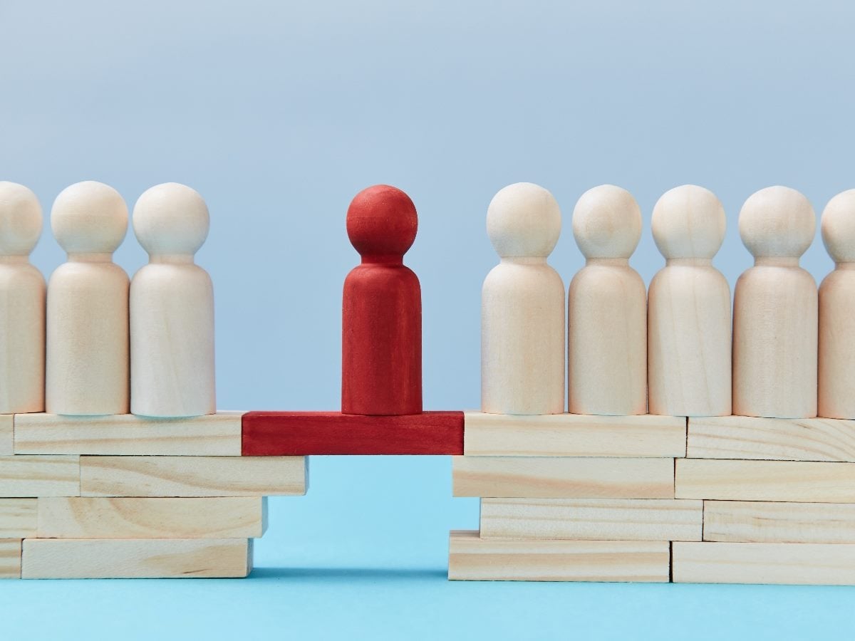 A line of light wooden figures stands on each side of a gap, connected by a red wooden bridge with a single red figure in the center, symbolizing a leader closing organizational or communication gaps.
