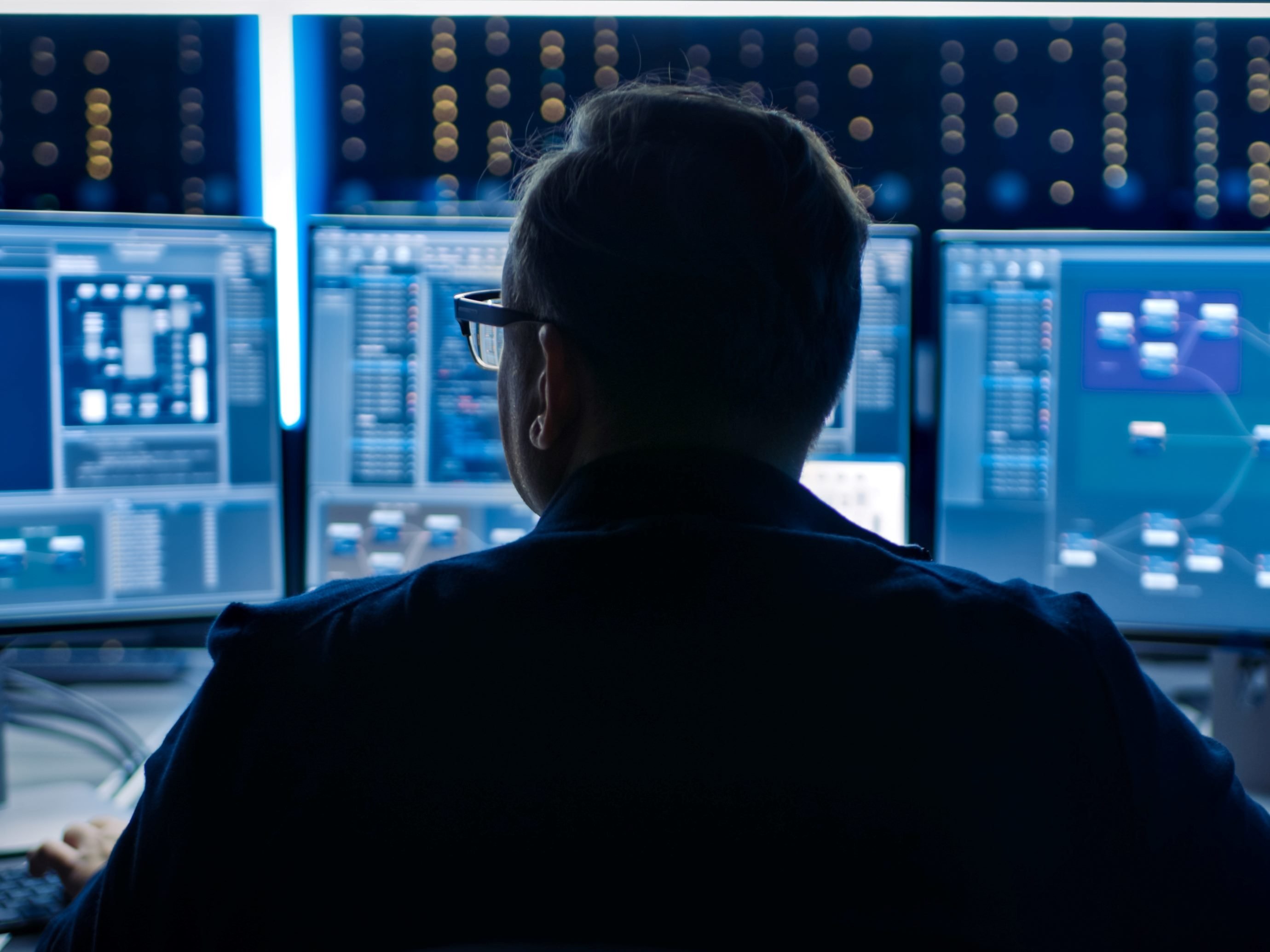 An IT professional. sits with back toward the screen working in a data center with three displays in front of them in piece about AI cybersecuity.