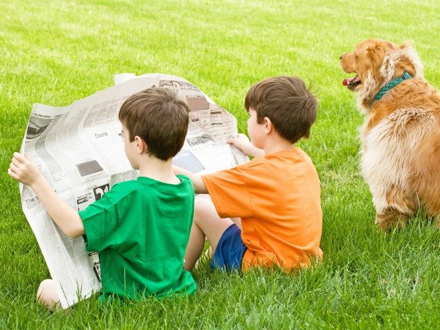 Two boys sitting in grass reading a newspaper next to a dog sitting in grass. The dog is not reading a newspaper.