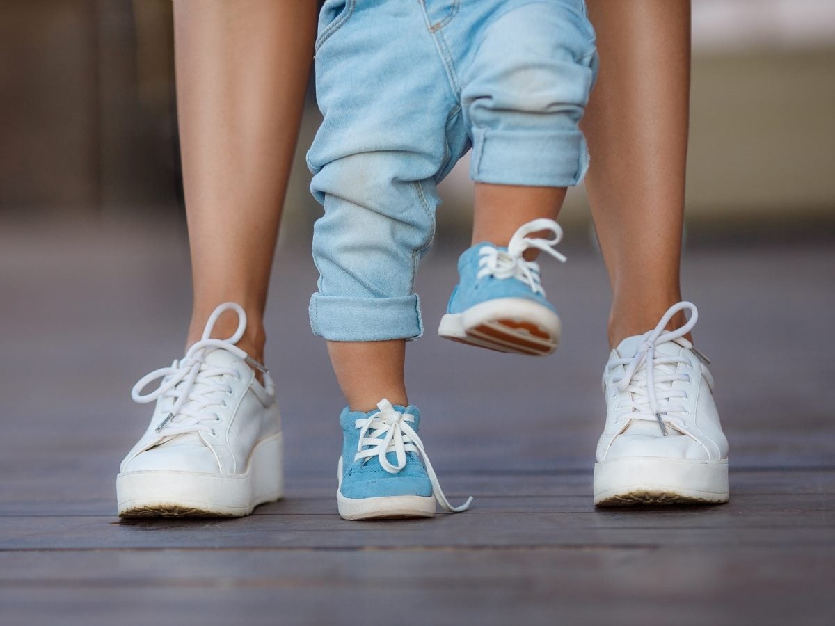 A toddler in cuffed light denim pants and blue suede sneakers takes a step between an adult's legs, one small foot lifted mid-stride in blue-and-white shoes while the other rests on a wooden deck, framed by the adult's white platform sneakers and bare legs in soft, warm outdoor light.