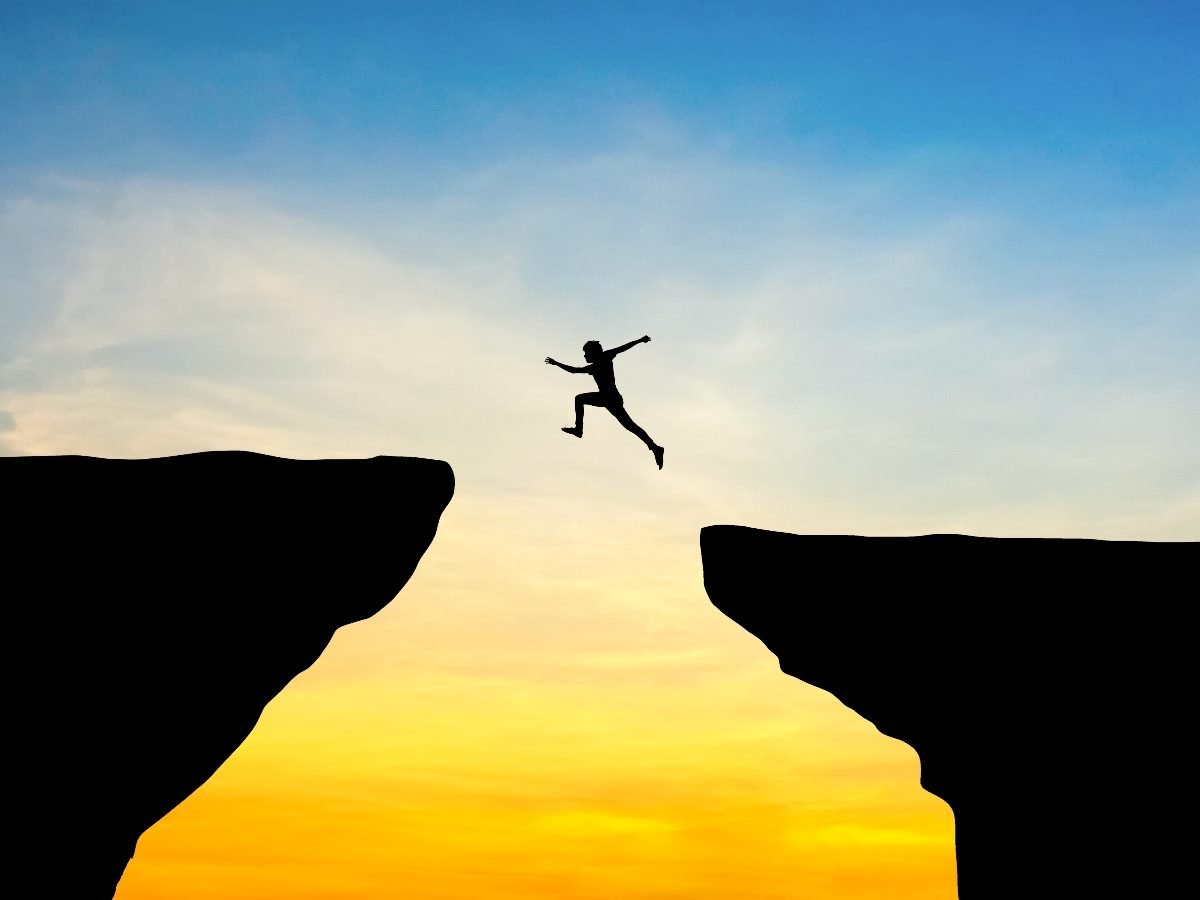Silhouette of a person leaping across a wide gap between two cliffs at sunset, symbolizing risk, challenge and bridging a difficult divide.