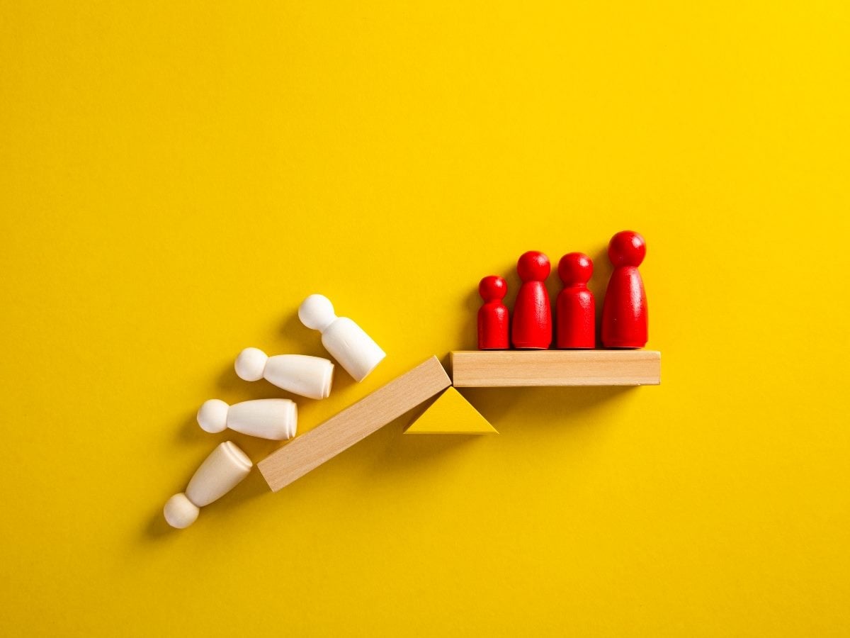 Wooden peg figures on a seesaw illustrate imbalance, with four red figures elevated on one side while several white figures fall off the lower side against a bright yellow background.