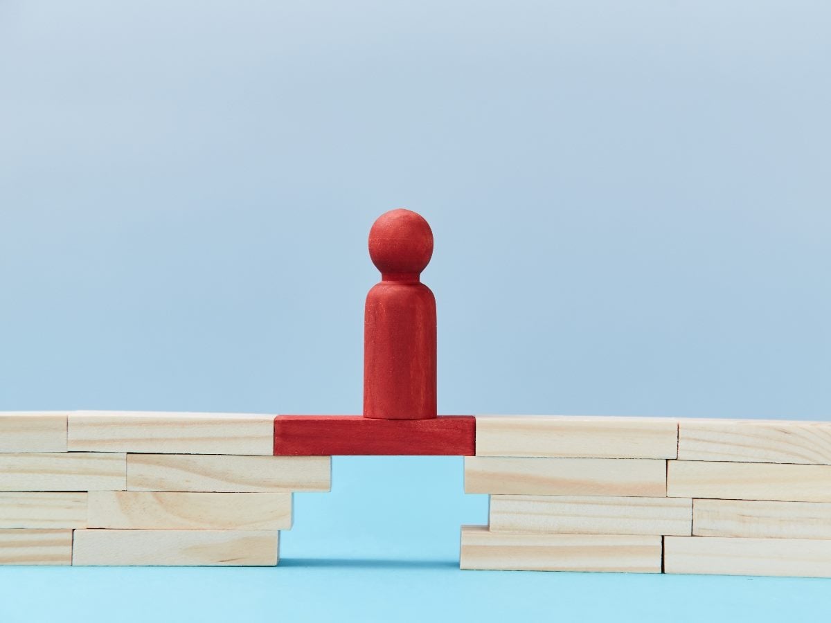 Wooden blocks form two uneven platforms separated by a gap, with a red game-piece figure standing on a single red plank bridging the opening, symbolizing connection, leadership, stability or closing operational gaps.