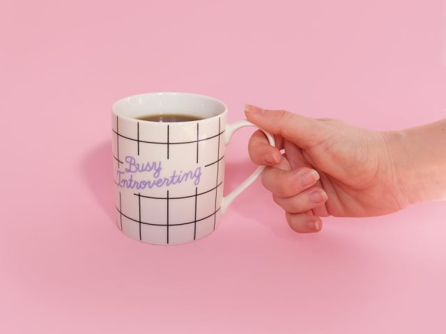 hand holding a white coffee mug with the message Busy Introverting against a pink background