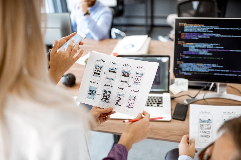 This image shows two professionals, possibly in a tech or design office setting, reviewing mobile app interface sketches on paper. The person in the foreground has blonde hair, holds a smartphone in one hand and a paper with app designs in the other, while the other person, looks on attentively. The backdrop includes a laptop displaying code and other work-related items, suggesting a collaborative workspace environment focused on product features and app development or user experience design.
