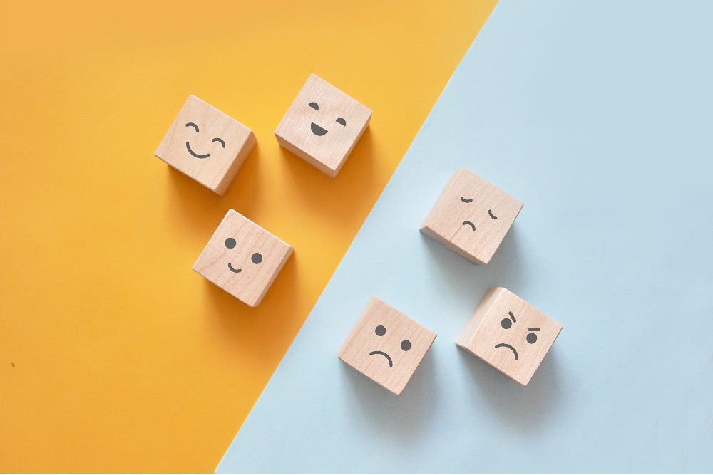 Faces drawn on six wooden cubes illustrate various human emotions against a yellow background for the happier emotions and a blue background for the sad or angry emotions in piece about using psychographics in marketing.