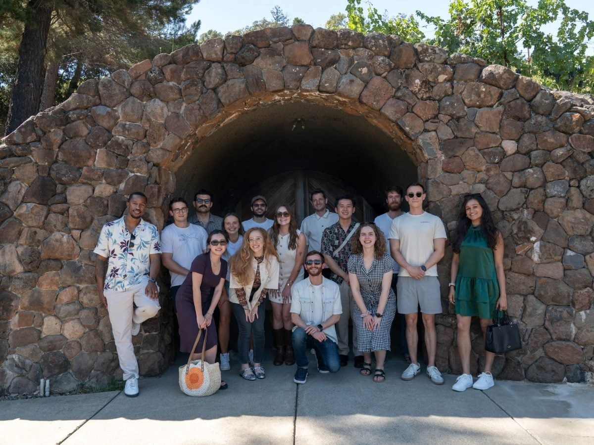 A group of more than 15 Outset team members smiling and standing together in front of a stone-arch wine cave during an offsite celebration. The photo reflects how the team has grown from 12 people at Series A to over 30 at Series B, showcasing the diverse, world-class group building the company’s AI-moderated research platform.