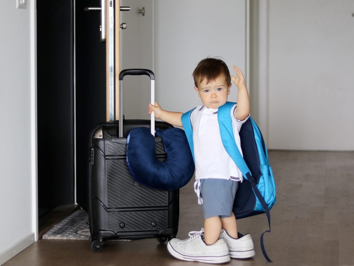 Toddler standing by an open doorway, holding the handle of a small rolling suitcase and waving, wearing a backpack, oversized sneakers, and a neck pillow as if ready to travel.
