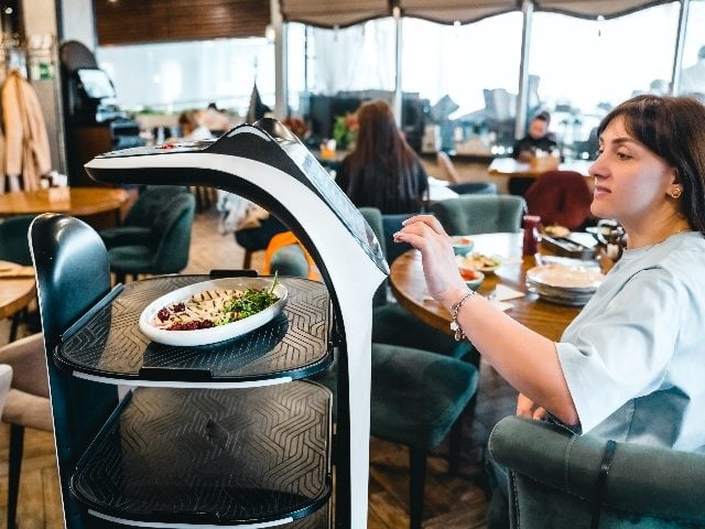 A customer at a restaurant sitting at a table ordering from a robot-like waiter machine.  