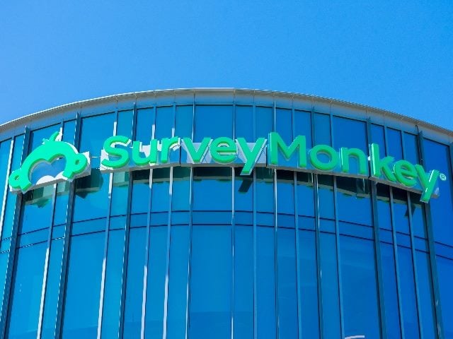 The SurveyMonkey name and logo in green against a modern steel and glass curved building with a blue sky in the background, in a story about the rebranding strategy of SurveyMonkey.