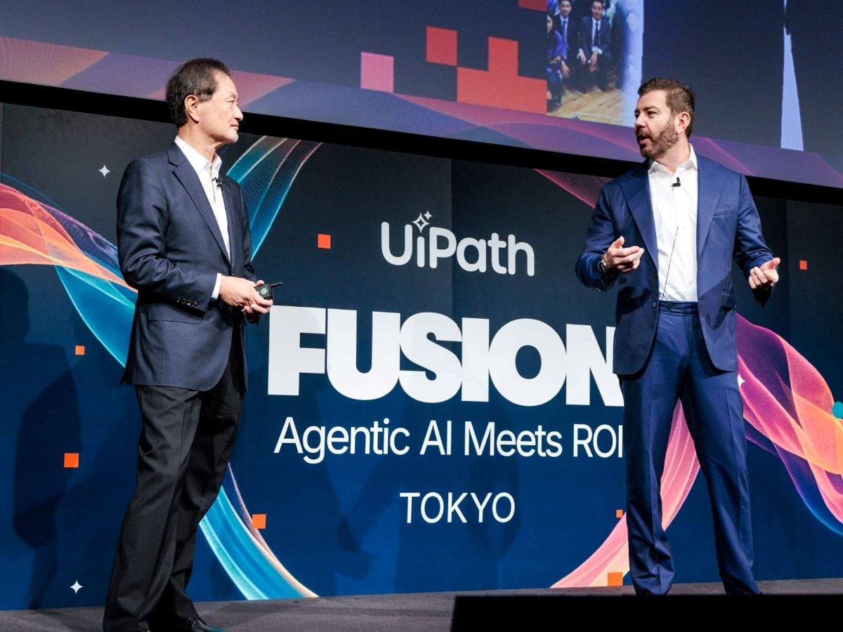 Two presenters stand on stage at the UiPath FUSION event in Tokyo, speaking in front of a large backdrop that reads “Agentic AI Meets ROI.” Both are dressed in business suits and appear engaged in discussion as they address the audience.