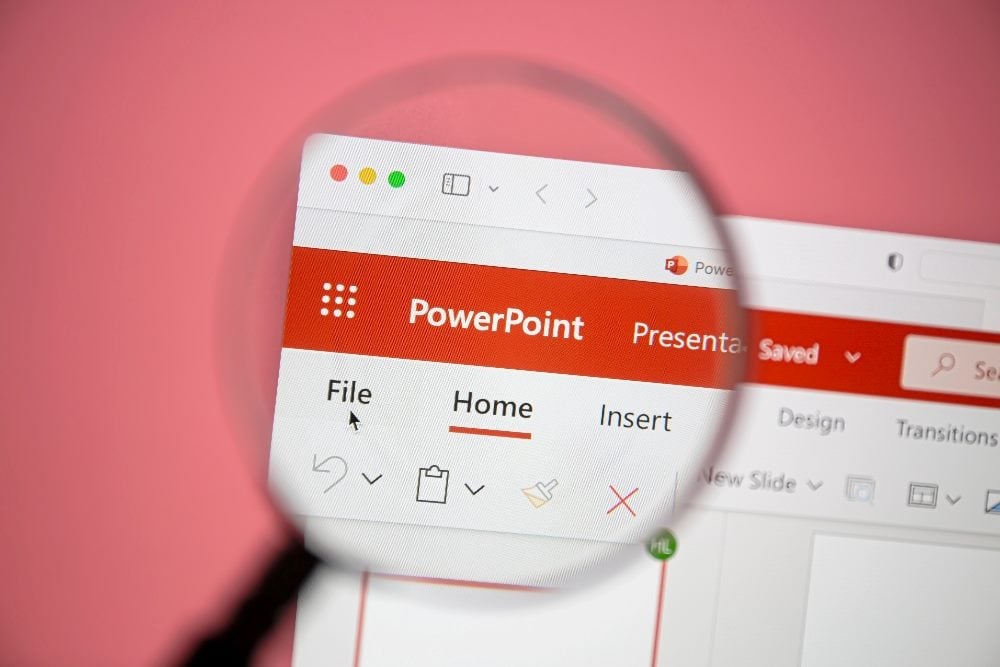 A close-up image of a computer screen displaying a PowerPoint application window, with the "Home" tab selected. The screen is magnified by a magnifying glass, highlighting the "File" menu and toolbar options. The background is a solid pink color, adding contrast to the white and red interface of the PowerPoint application in piece about ditching PowerPoint in Customer Advisory Board Meetings.