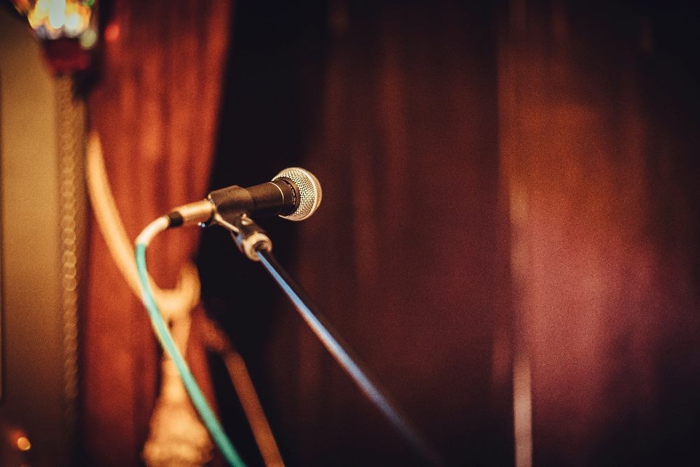 The image shows a microphone positioned on a stage, bathed in warm, ambient lighting. The backdrop features curtains, contributing to an intimate and focused setting, suggestive of a live performance or speech. The microphone, as the central focal point, stands ready to amplify the voice of the next speaker or performer, symbolizing communication and expression in piece about brand voice.