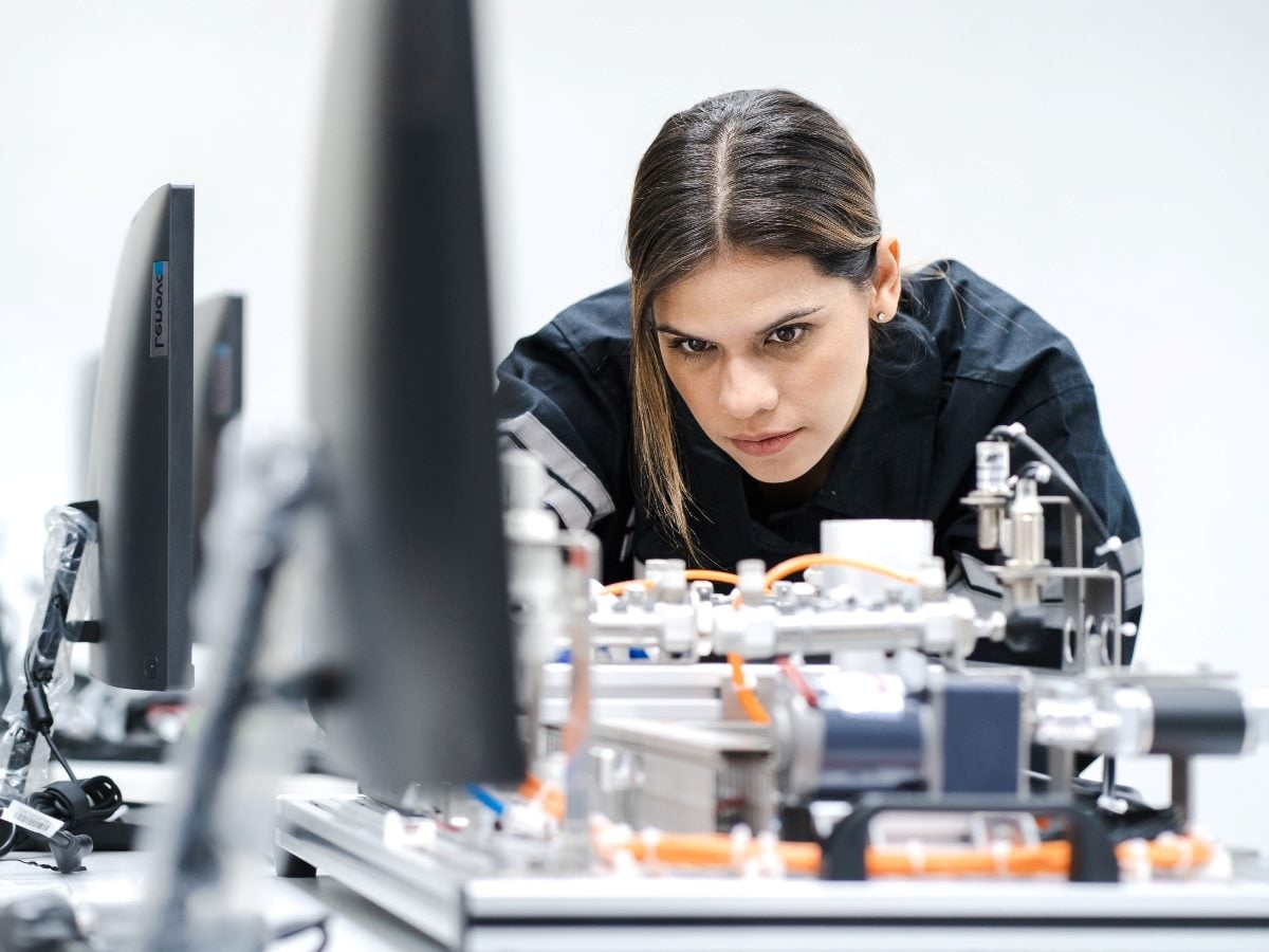 Person concentrating while working with industrial equipment and machinery in a bright technical workspace, with computer monitors and engineering components visible in the foreground.