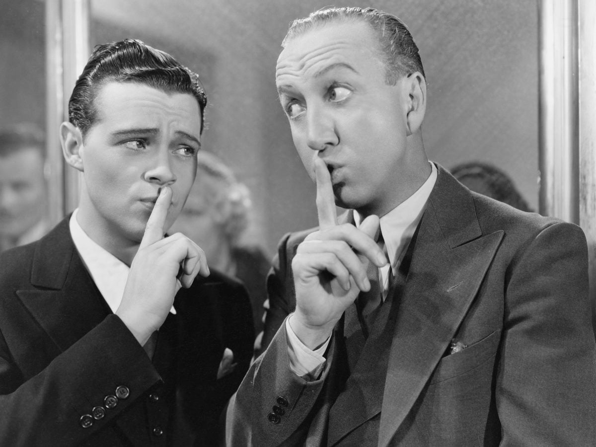 Black-and-white vintage photograph of two men in suits standing close together indoors, each holding a finger to his lips in a "shh" gesture. Both appear to be exchanging a conspiratorial glance, suggesting secrecy, discretion or silence, while blurred figures stand in the background.