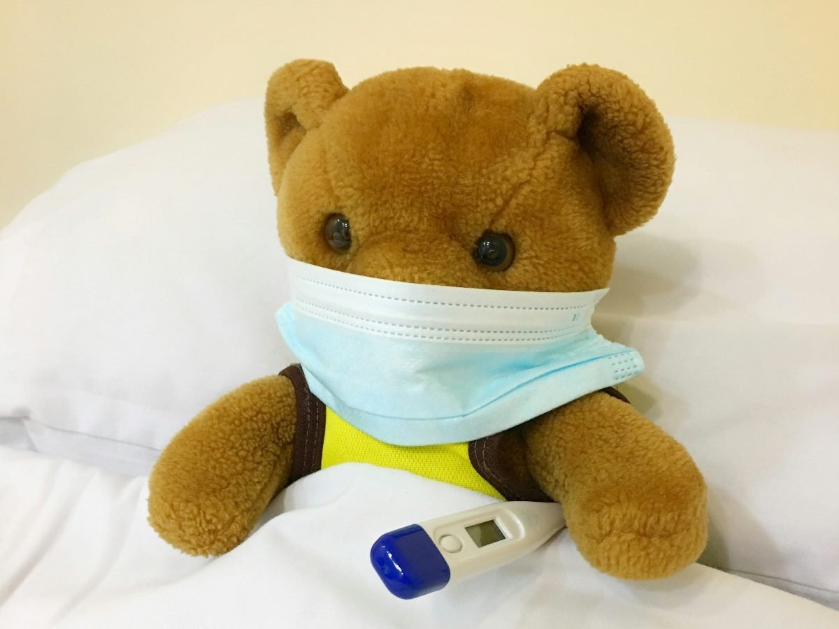 Teddy Bear with a health mask on holding a thermometer while laying in bed.