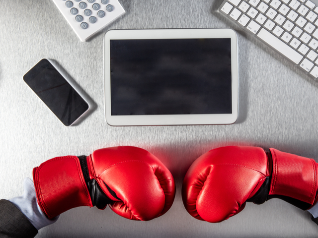 Boxing gloves facing technology screens, digital tablet, calculator and computer on an office desk - project management tool showdown