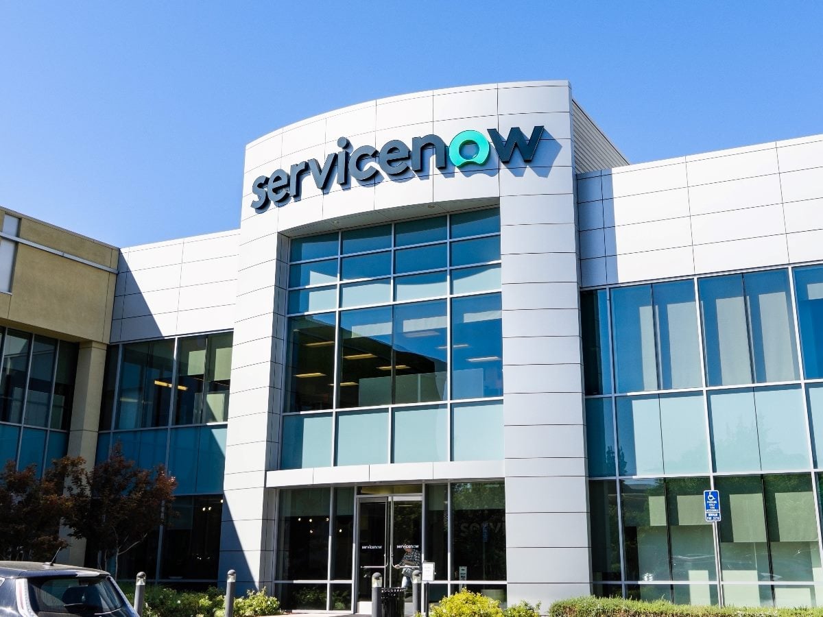 Exterior view of a modern office building with large glass windows and a curved facade displaying the ServiceNow logo above the main entrance under a clear blue sky.