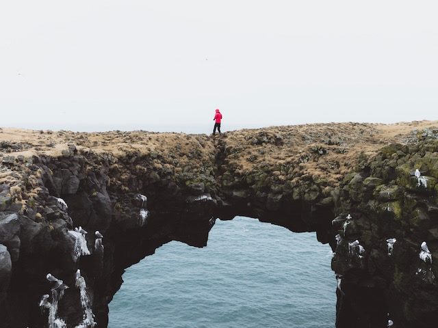 person in red parka walking over natural bridge over water