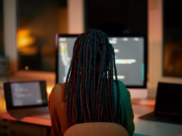 Back view portrait of female IT developer writing code on multiple computer screens while working late in dark office. 