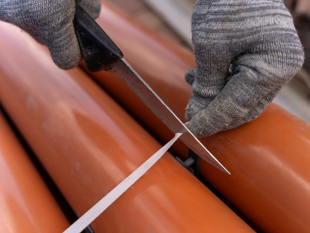 A gloved hand grips a knife with a black handle to cut a strap of white plastic packing tape, positioned in a close-up over rows of copper-colored pipes or tubing bundled together outdoors.