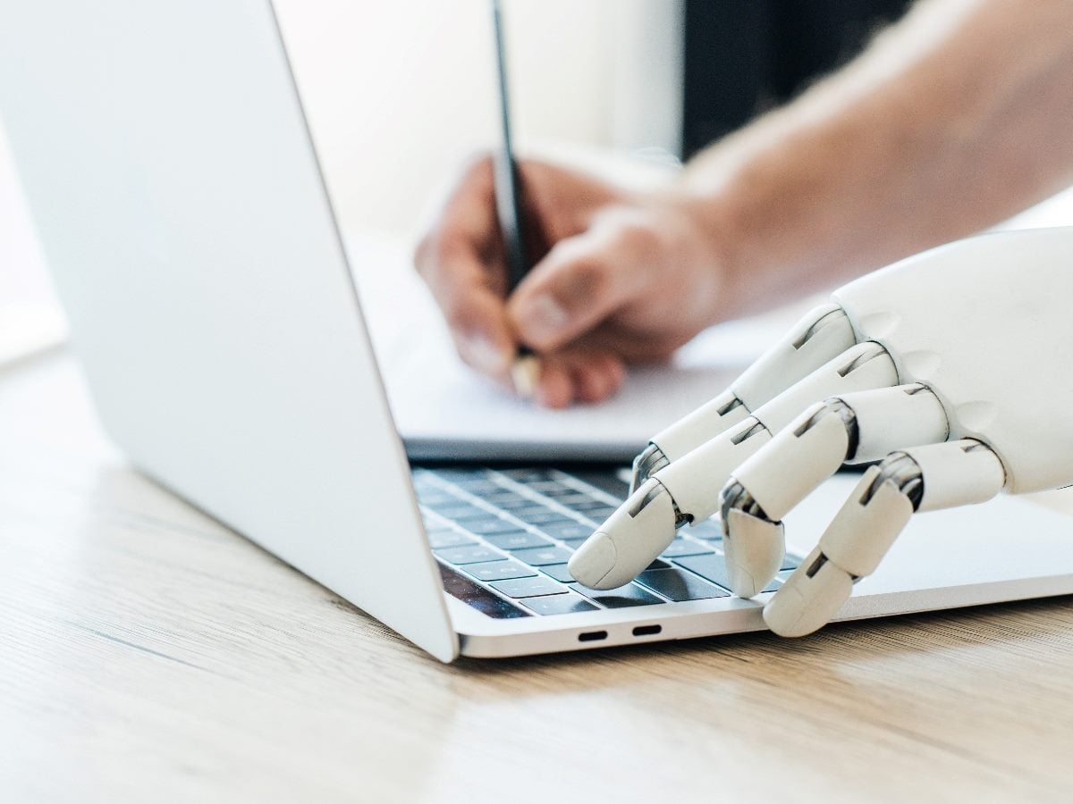 Close-up image showing a robotic hand using a laptop keyboard beside a human hand writing on paper, illustrating collaboration between AI systems and human work in a digital environment.