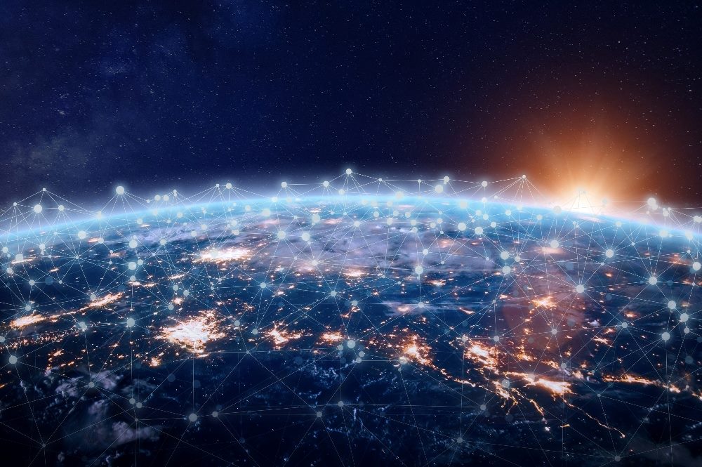 The image depicts a view of Earth from space, illuminated by city lights at night, overlaid with a glowing digital network of connected nodes and lines. The nodes and lines create a web of connections that envelop the planet, symbolizing global communication and data networks. The horizon glows brightly with a sunset or sunrise, adding a dynamic contrast between the dark space and the lit Earth in piece about customer data and the connected world.