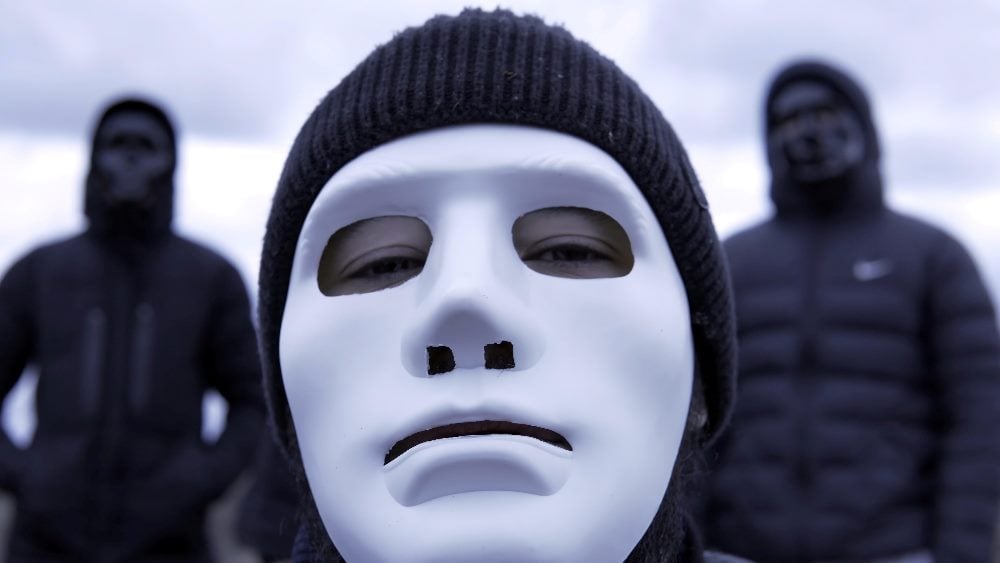 Criminal gang in black plastic masks and leader in white mask on background of cloudy sky in piece about fake podcast scam.