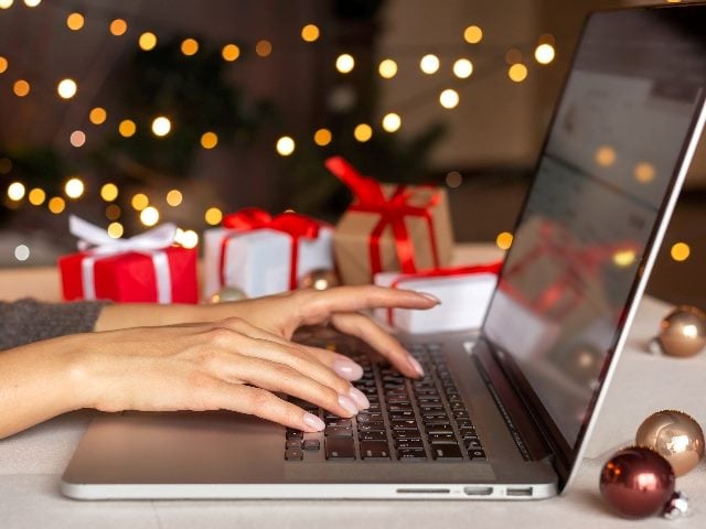 A person with manicured nails types on a laptop with strings of holiday lots and small cheerfully wrapped gifts in the background in piece about this holiday shopping season trends. 