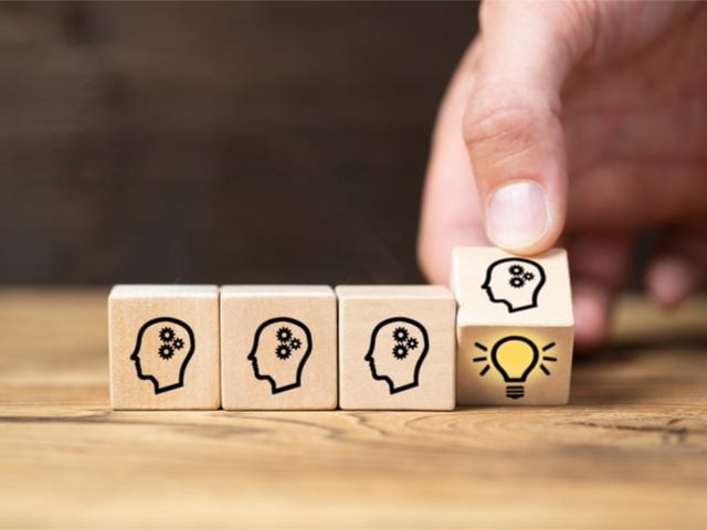 Three dice with hand drawn heads on them. A hand is half turning one over and there is a lit light bulb on the bottom - innovation concept