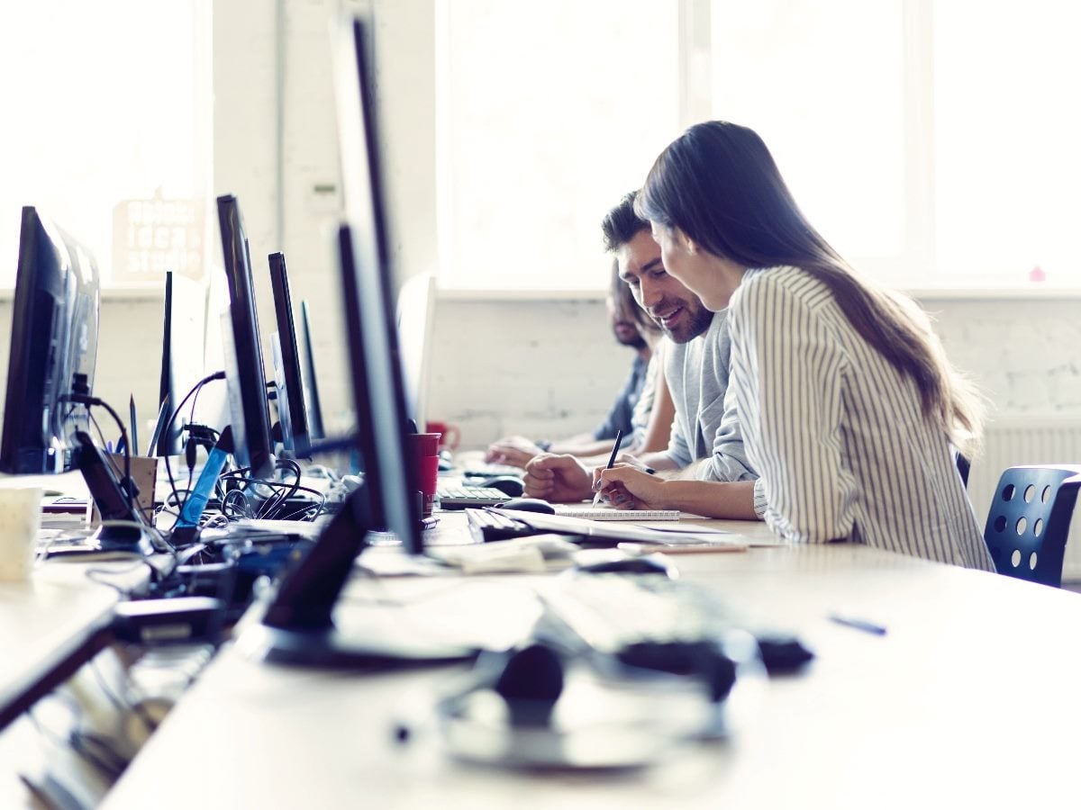 Customer service team members working at computers in an office contact center, collaborating while reviewing notes at their desks.