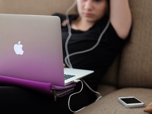 woman sitting on couch looking at her laptop with earphones in, cellphone next to her