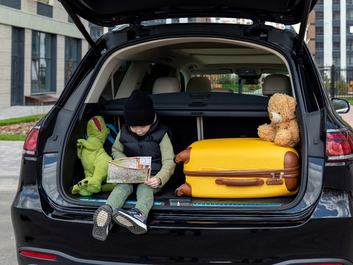 A young child sits in the open trunk of a car, holding a map while seated between a green dinosaur plush toy and a yellow suitcase with a teddy bear resting on top.