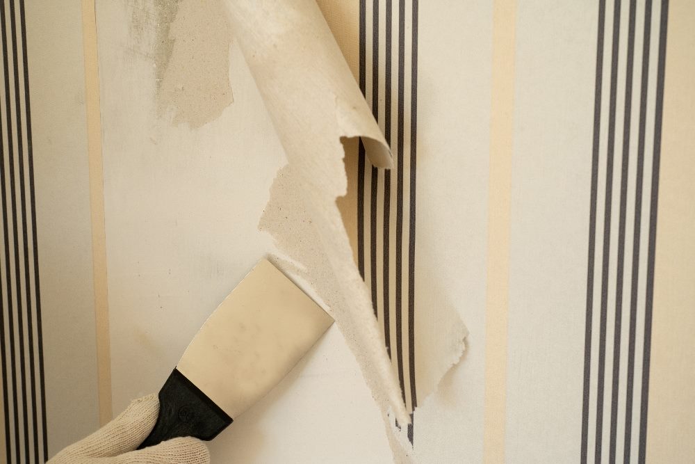 A hand in a white glove is using a putty knife to peel back old wallpaper, revealing the bare wall underneath. This image symbolizes the process of adopting a new digital experience platform and the importance of thorough change management to ensure a smooth transition and long-term success.