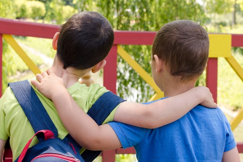 Two kids wearing T-shirts and one wearing a backpack stand together with their arms on each others' shoulders in front of a red and yellow painted rail with trees in the background in piece about CFO and marketing alignment.