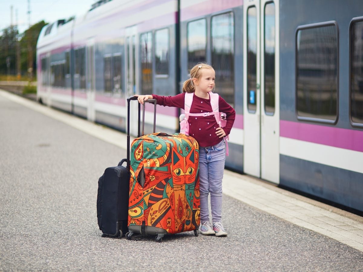 A young girl with a blonde ponytail stands on a train platform beside a large suitcase decorated with a colorful cat design in teal, orange and yellow, resting one hand on its extended handle. She wears a maroon long-sleeve top with a small heart print, light-wash jeans, sneakers and a pink backpack, and looks off to the side with one hand on her hip. A smaller black suitcase sits next to the patterned one, and a purple-and-white train with its doors open stretches along the platform behind her.