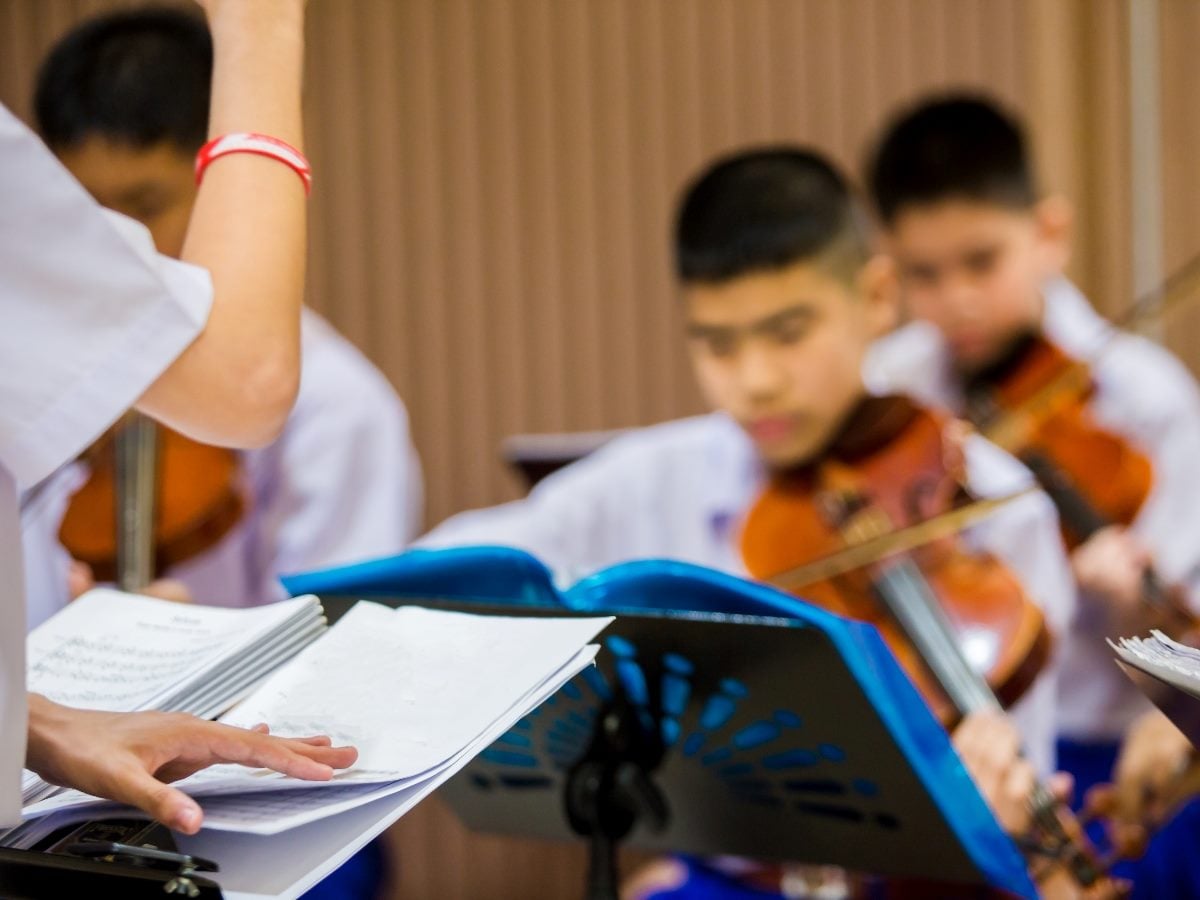 A conductor holding sheet music leads a group of young violinists performing together, with music stands and instruments in a rehearsal setting.