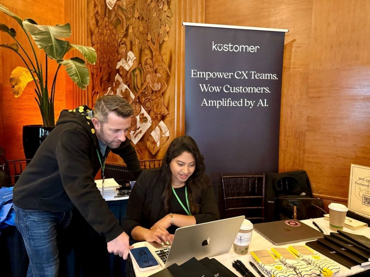 Two Kustomer team members collaborate at a laptop during an event, seated at a booth with a banner reading “Empower CX Teams. Wow Customers. Amplified by AI,” surrounded by branded materials and conference items.