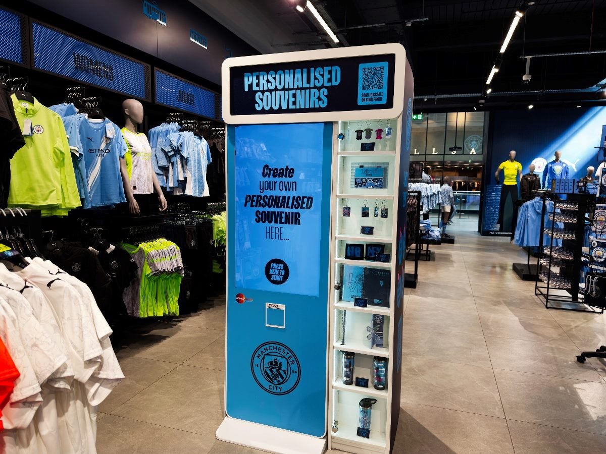 A retail display inside the Manchester City store features a blue “Personalised Souvenirs” kiosk inviting customers to create their own customized merchandise. Surrounding the kiosk are racks of Manchester City apparel, including jerseys, T-shirts and sportswear in team colors.