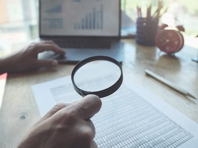 Business person using magnifying glass and laptop computer to audit and check the  integrity of information sheets in piece about CX audits. 