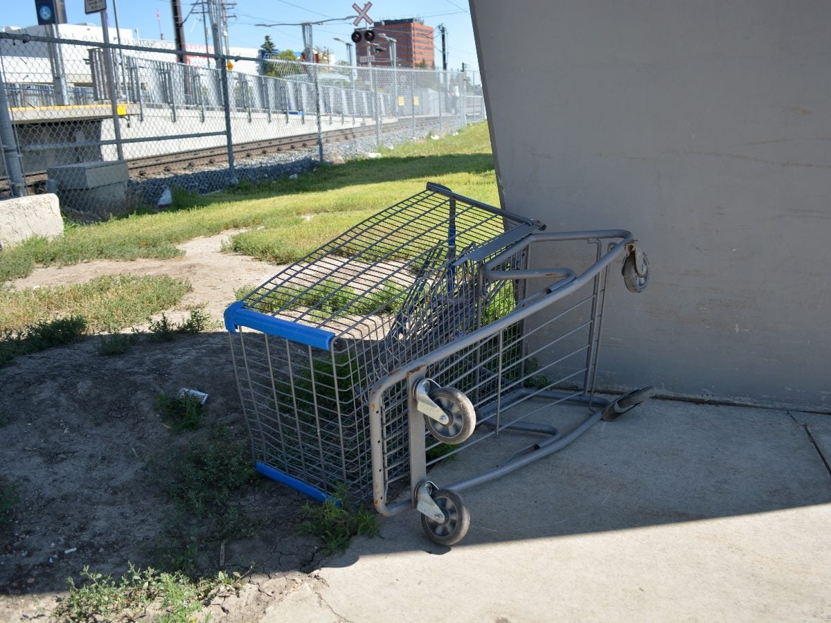 Abandoned shopping cart tipped on its side beneath a concrete overpass near train tracks, with fenced rail lines and urban buildings visible in the background on a sunny day.